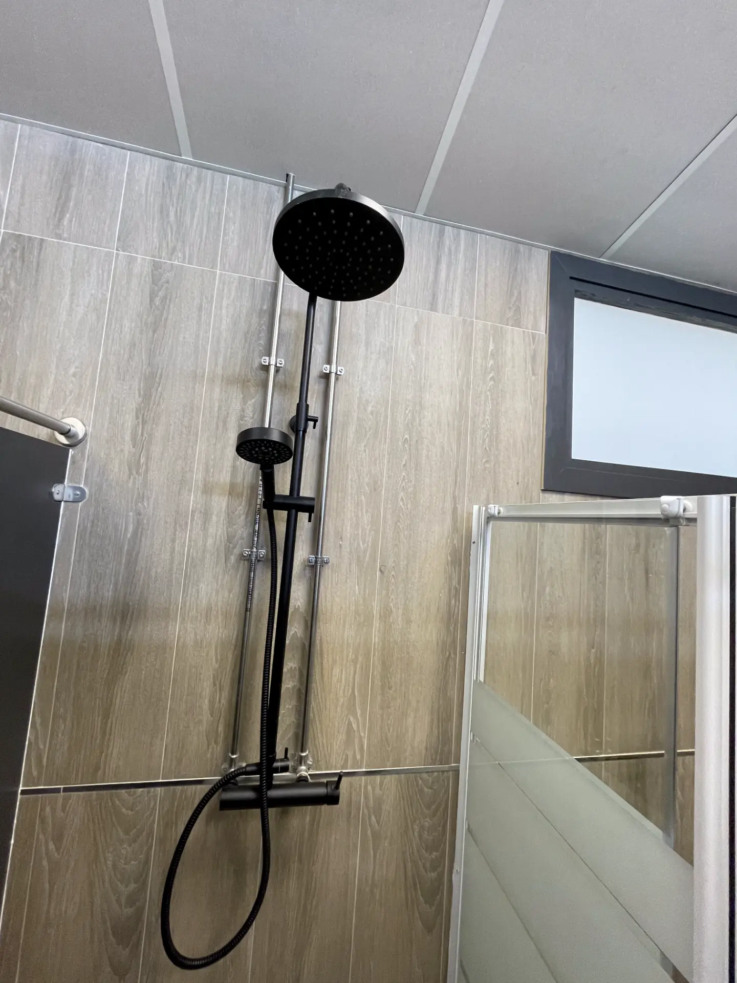 Columna de ducha termostática negra instalada en un baño con pared imitación madera